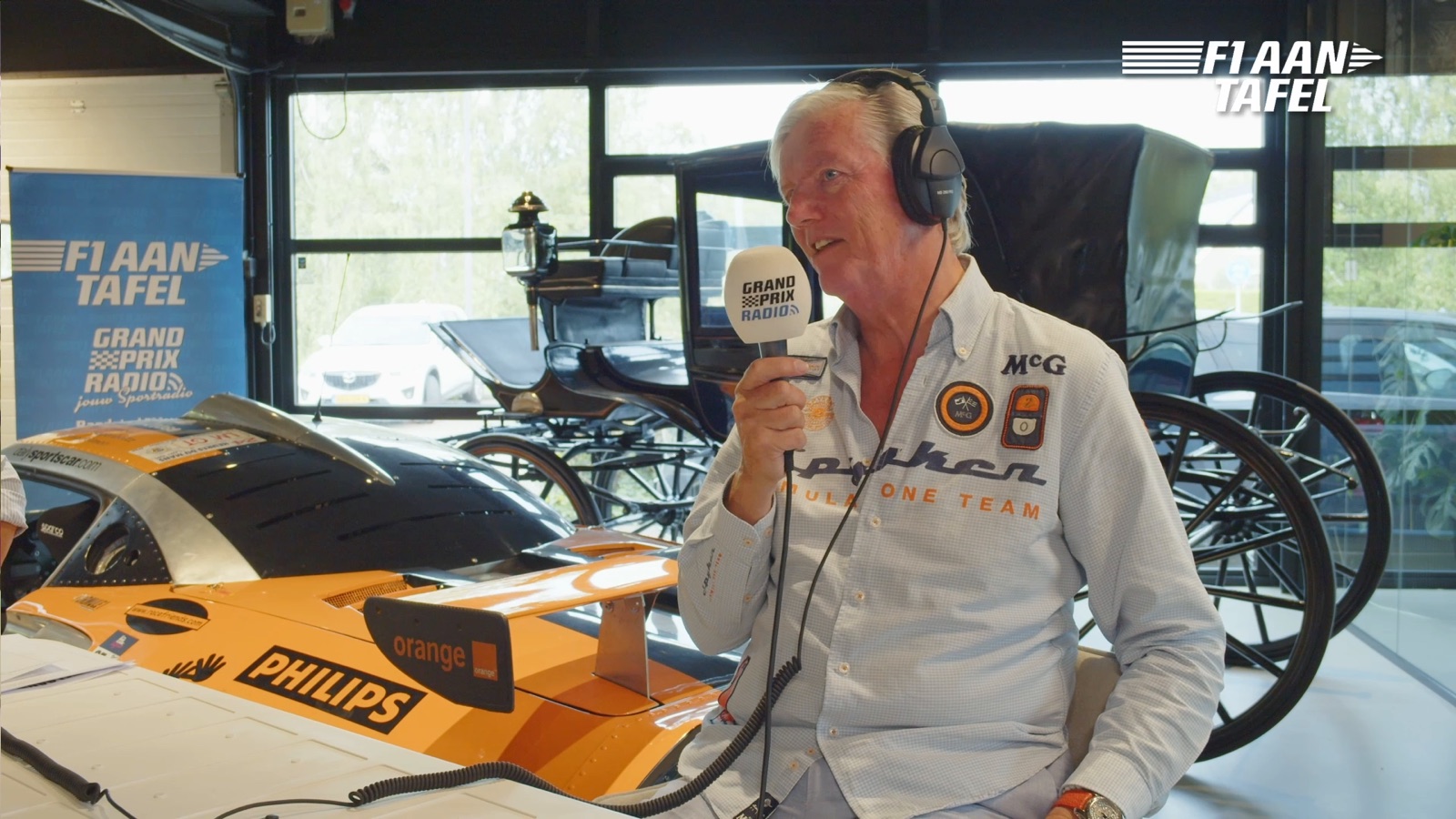 Presentator met microfoon naast een oranje Spyker-racewagen tijdens een special van F1 aan Tafel