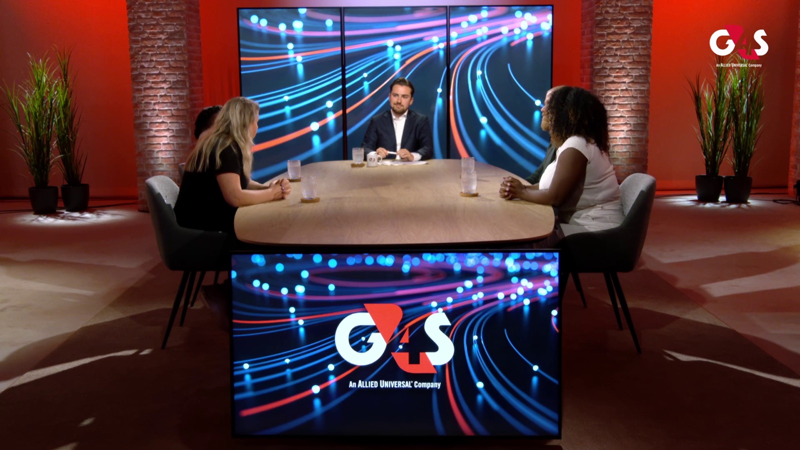 Brede opname van de talkshowset van G4S: presentator en gasten aan tafel, logo op de schermen